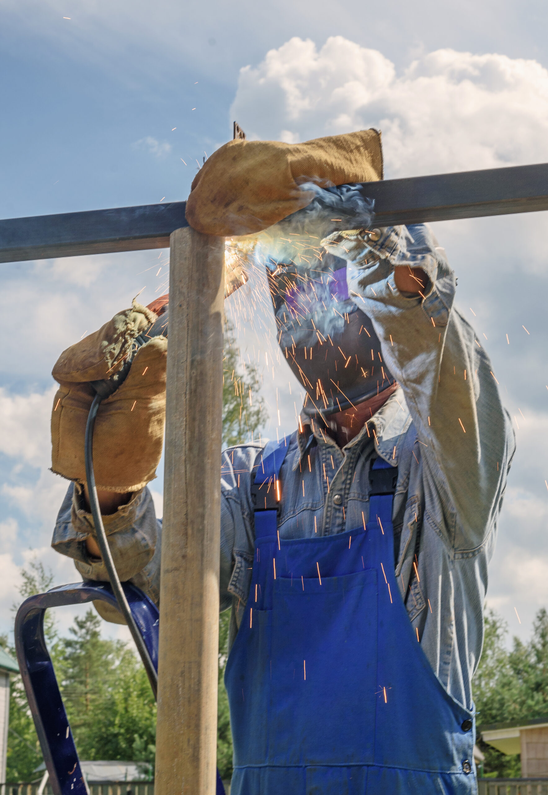 Man welder in a welding mask, construction uniform and protective gloves cooks metal on a street construction site. Construction of a pavilion, pergola near a country house on a summer day. Excavator bucket dropping dirt into an open trench.